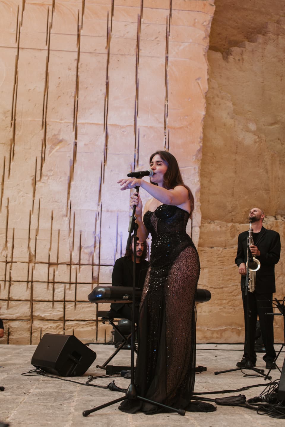 Maya cantando con un vestido de noche negro, con un teclista y un saxofonista detrás